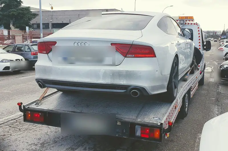Grúa particular con una berlina blanca cargada en un aparcamiento lleno de coches