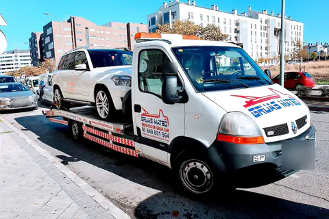 Grúa particular con un todocamino blanco cargado en una calle con edificios de viviendas